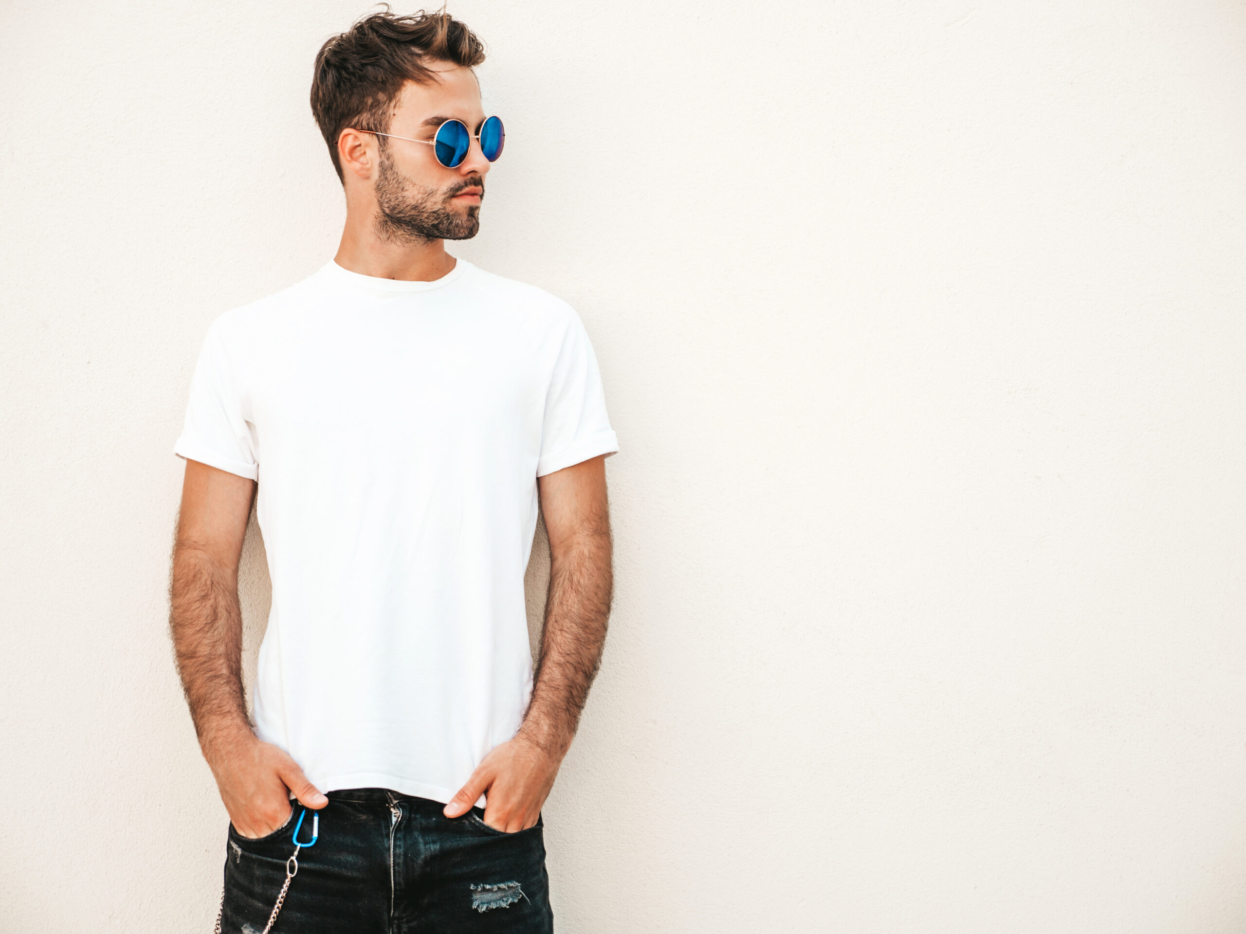 Portrait of handsome confident stylish hipster lambersexual model.Man dressed in white T-shirt. Fashion male posing on the street background near wall in sunglasses outdoors
